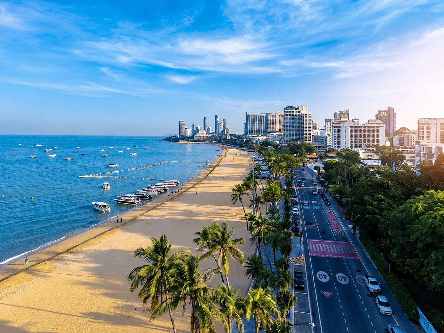 Beach Road, Pattaya จากมุมสูง: อ่าวโค้งรูปพระจันทร์เสี้ยว เรือเร็วจอดอยู่หน้าหาด ถนนเลียบชายหาดที่มีต้นปาล์มเรียงราย และตึกโรงแรมกับคอนโดมิเนียมของใจกลาง Pattaya อยู่ด้านหลัง