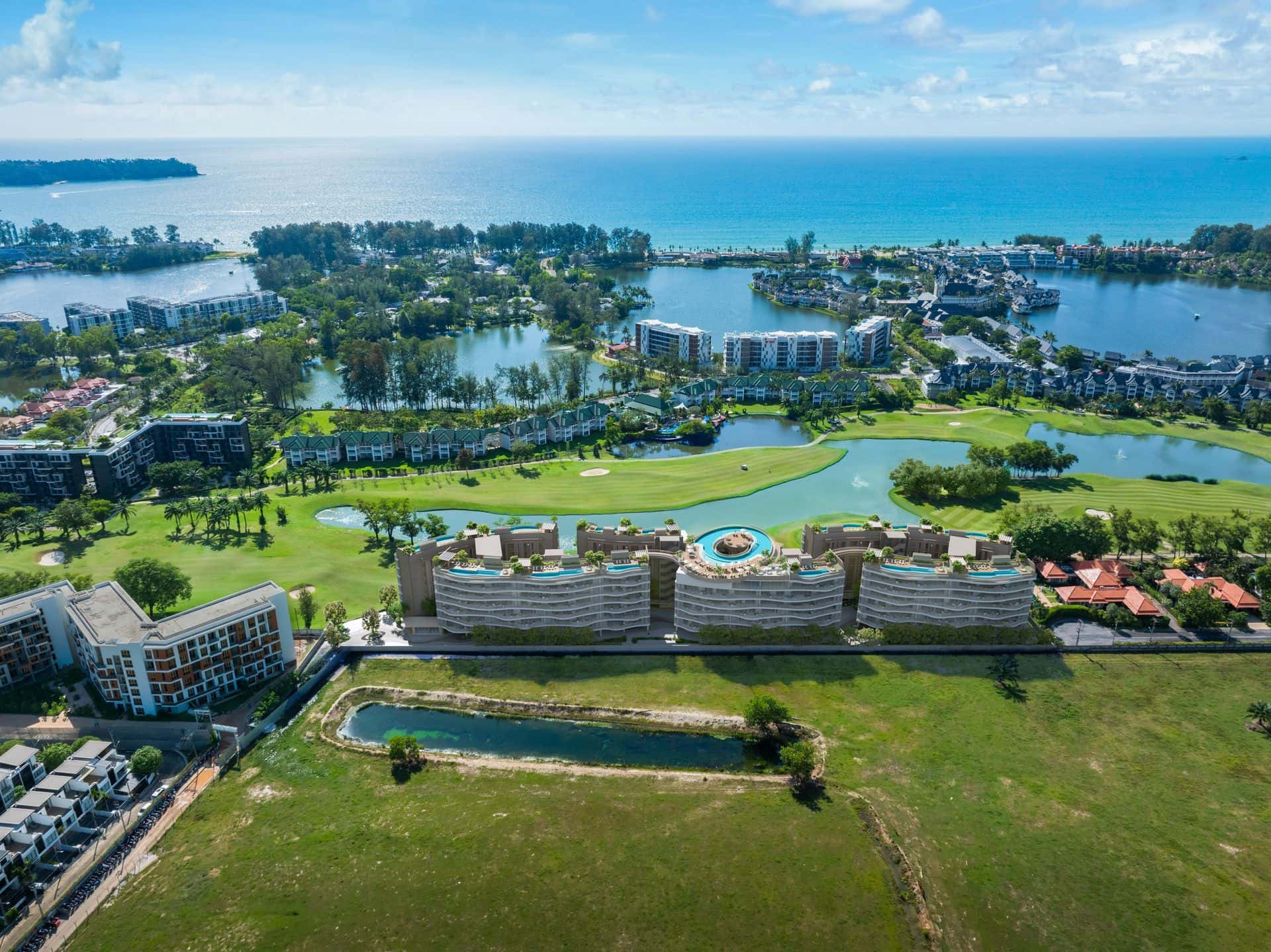 Laguna Phuket Angsana Golf Residences Topaz 的鳥瞰圖:三座弧形低層大樓與環形空中泳池位於高爾夫球場旁,遠處是潟湖與 Bang Tao 海灣。發展商的無人機實景相片,大樓以電腦合成加入