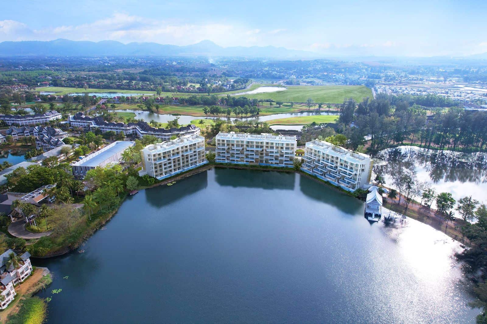 Aerial view of the three buildings of Angsana Oceanview Residences around the lagoon at the centre of Laguna Phuket, the golf course and the hills beyond: a render set into the developer's drone photograph, 2023