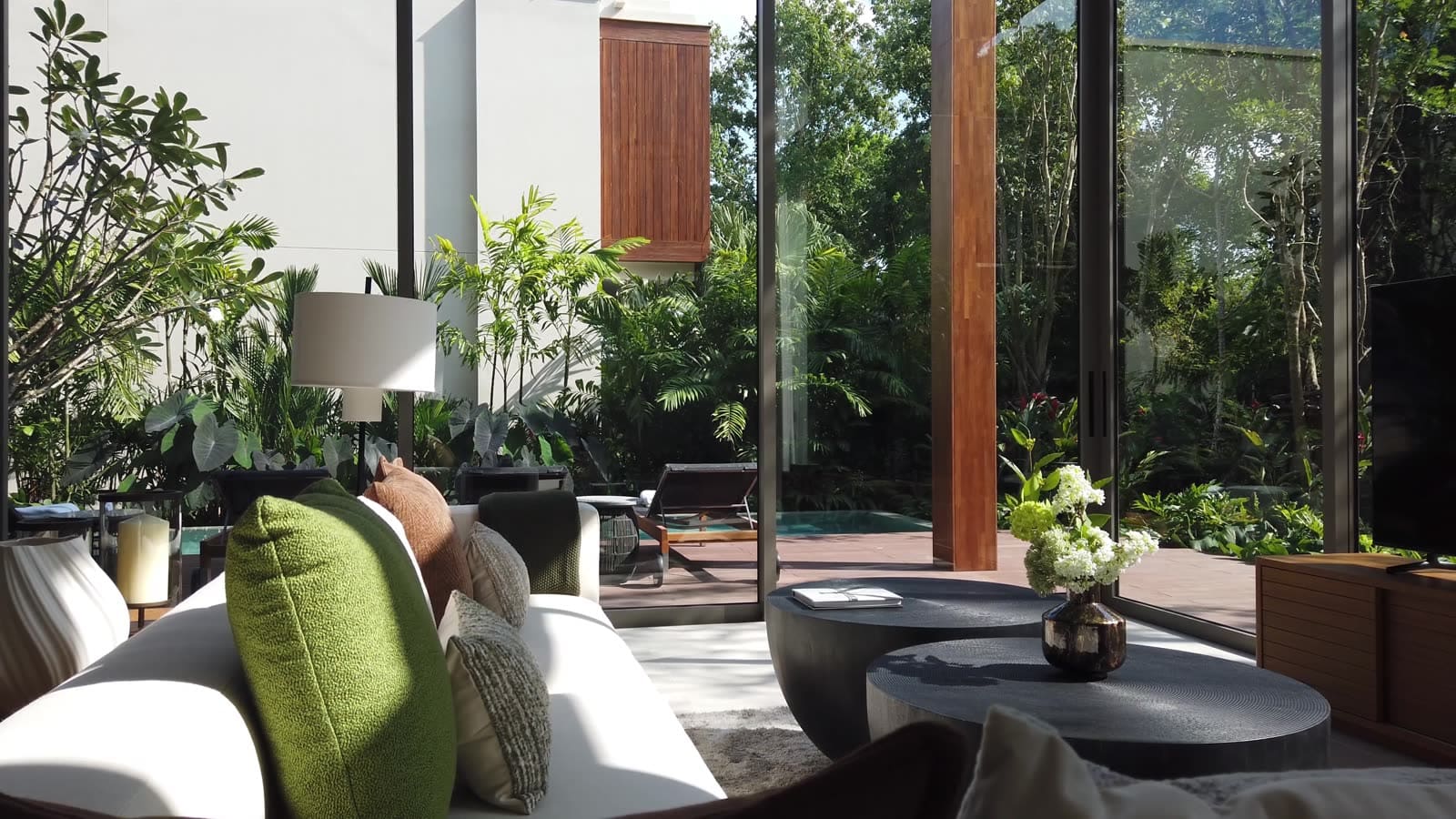 ห้องนั่งเล่นของวิลลาตัวอย่าง 4 ห้องนอนที่ Kiara Reserve Residences ภูเก็ต มองผ่านกระจกสูงจรดเพดานออกไปยังเด็คริมสระและสวน