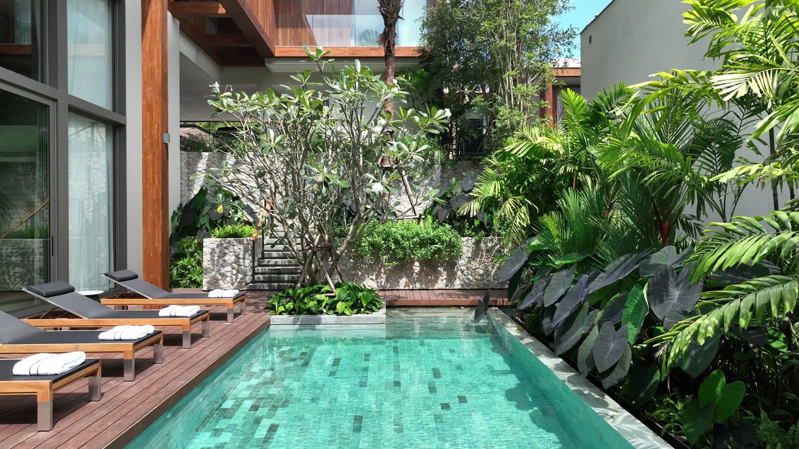 สระว่ายน้ำส่วนตัวของวิลลาตัวอย่าง 4 ห้องนอนที่ Kiara Reserve Residences อ่าว Layan ภูเก็ต กระเบื้องสีฟ้าคราม เด็คไม้พร้อมเก้าอี้อาบแดด ล้อมด้วยสวนแบบป่าเขตร้อน