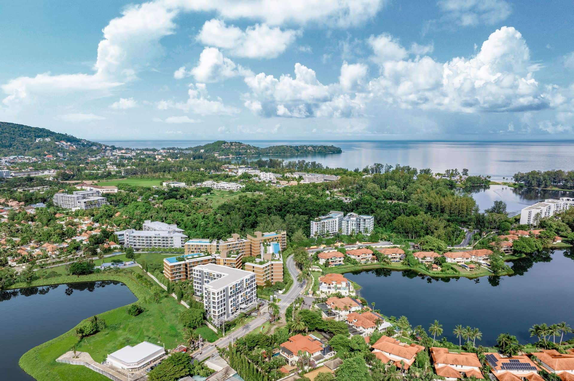 ภาพมุมสูงของ Laguna Lake Residences Aster ที่ทางเข้าหลักของ Laguna Phuket: ภาพถ่ายโดรนของผู้พัฒนาที่ใส่อาคารสามหลังเป็นภาพจำลองไว้ท่ามกลางทะเลสาบ มีอ่าว Bang Tao อยู่ไกลออกไป