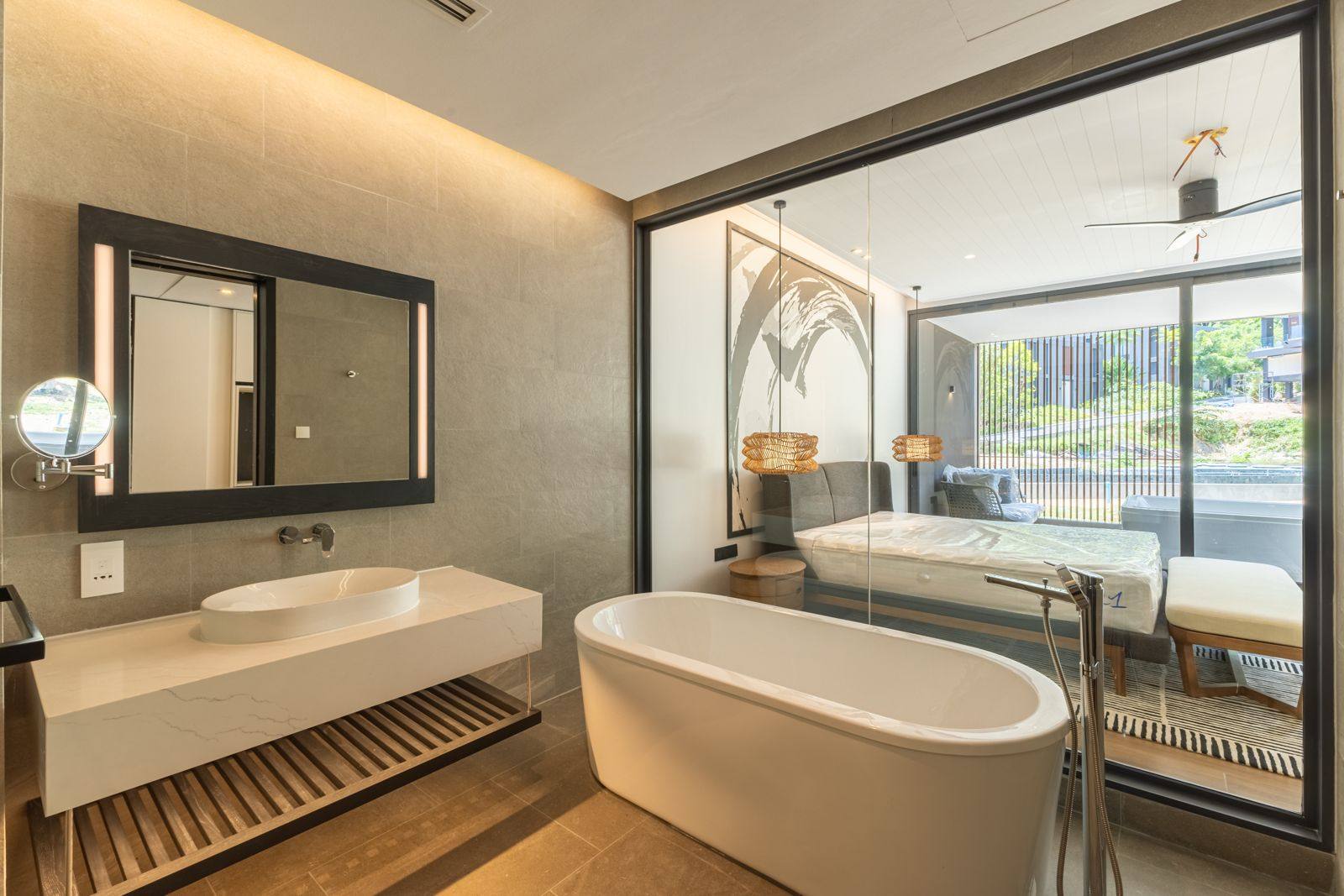 Ванная комната готовой квартиры-образца в MGallery Residences MontAzure Lakeside, Kamala, Пхукет: подвесная каменная тумба, накладная раковина, зеркало с подсветкой и отдельно стоящая ванна, за стеклом спальня