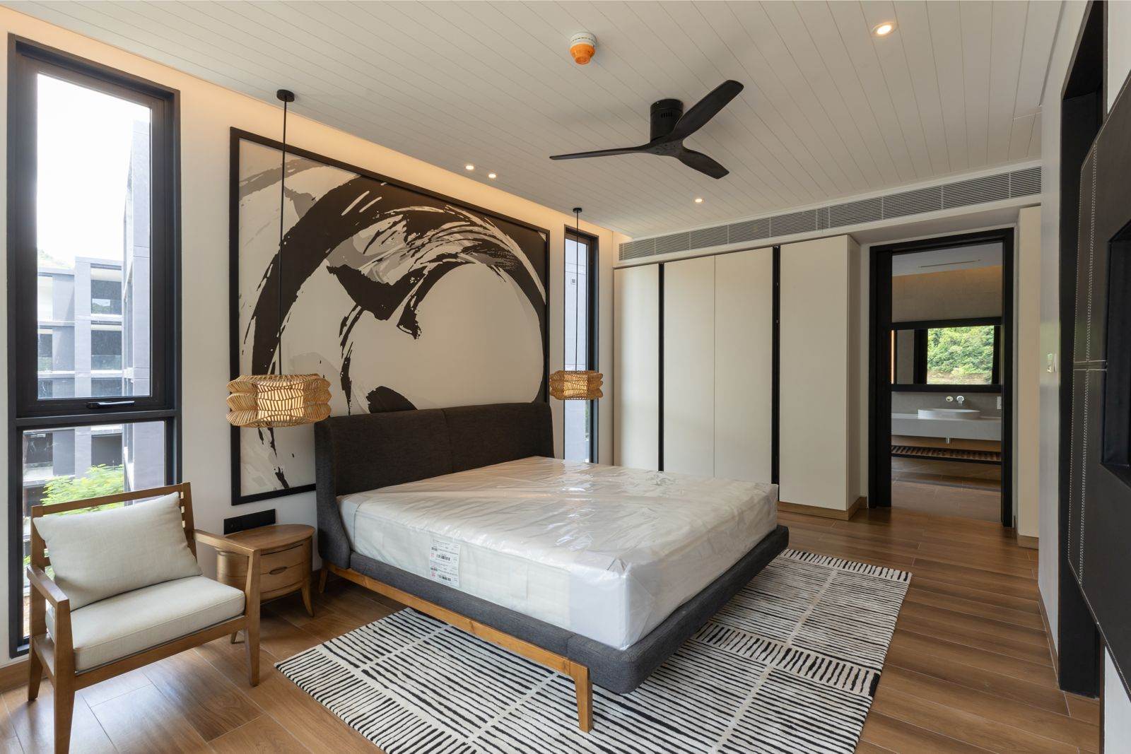 Спальня готовой квартиры-образца с одной спальней в MGallery Residences MontAzure Lakeside, Kamala, Пхукет: кровать под большой чёрно-белой картиной, ротанговые светильники и кресло