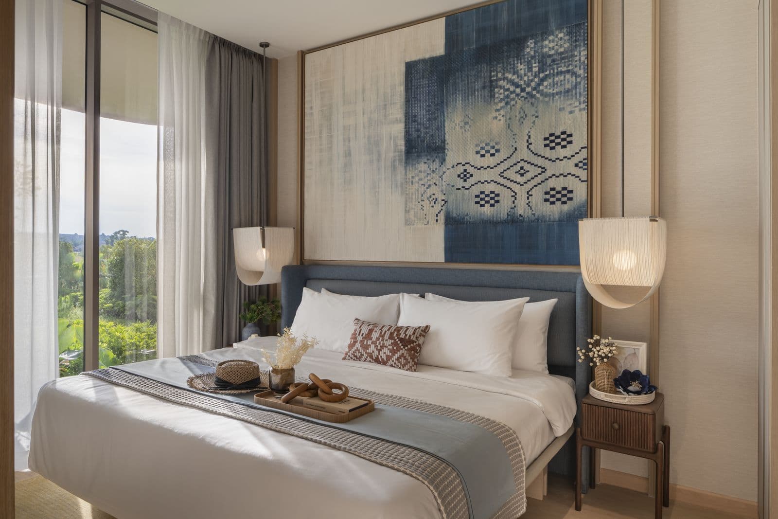 The bedroom of the finished one-bedroom show unit at PEYLAA Phuket: a king-size bed, a woven wall hanging and a floor-to-ceiling window onto trees