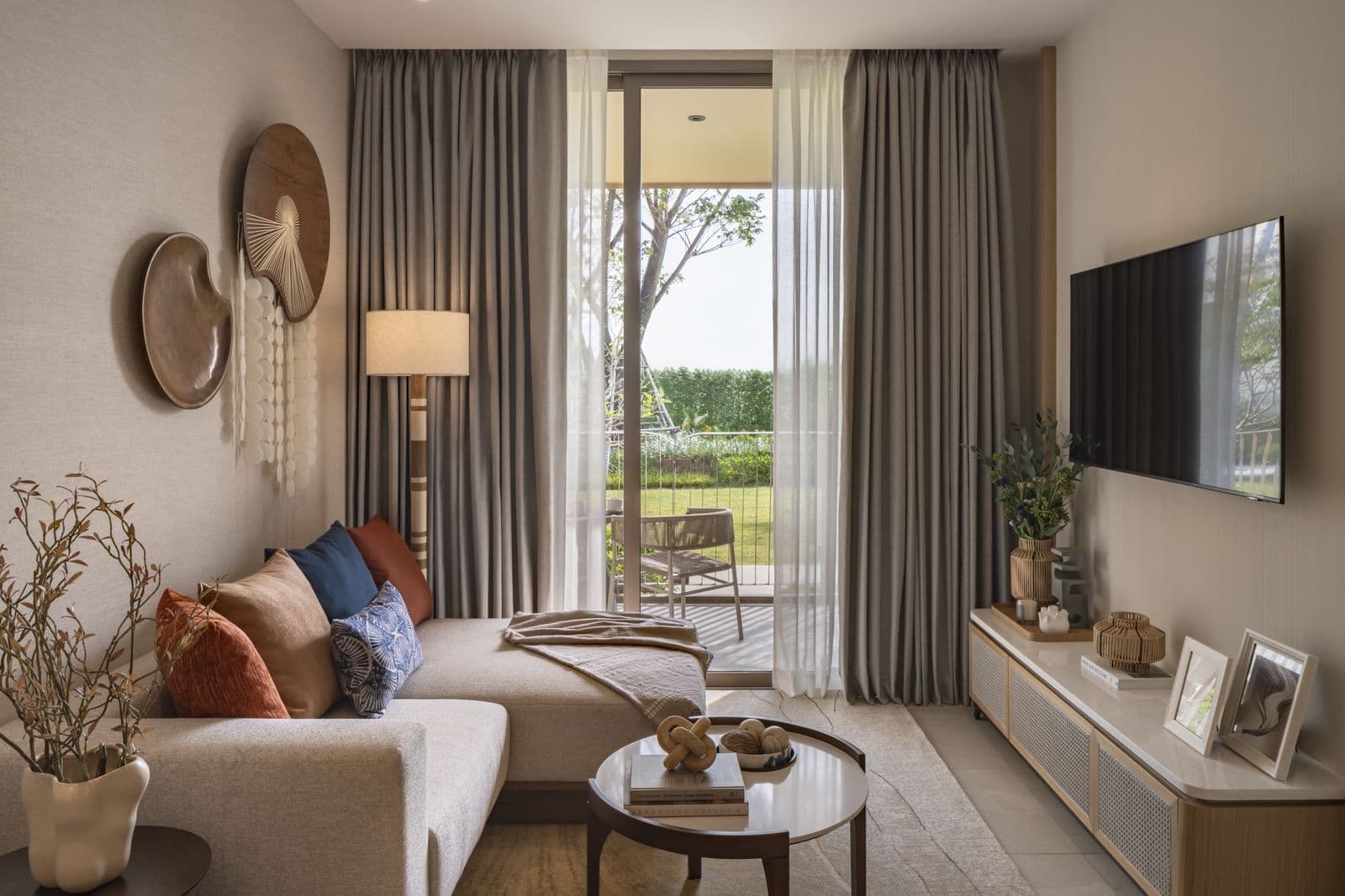 The living room of the finished one-bedroom show unit at PEYLAA Phuket: an L-shaped sofa, a round coffee table, a wall-mounted TV and a glass door to the balcony and garden