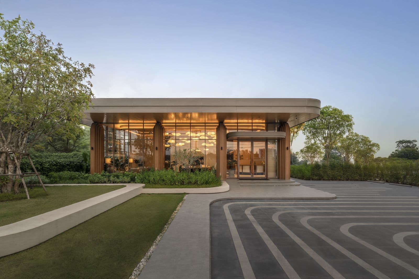 The PEYLAA Phuket sales gallery in Bang Tao: a single-storey glass and timber pavilion with a curved roof, lawn and a patterned driveway