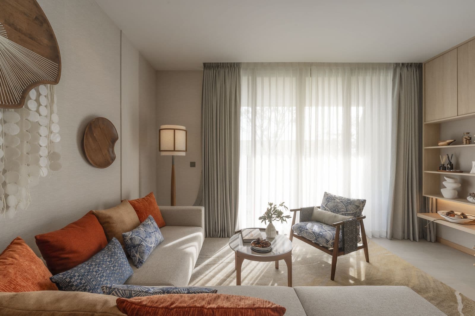 The living room of the finished two-bedroom show unit at PEYLAA Phuket: a sofa with orange and blue cushions, an armchair and tall curtained windows