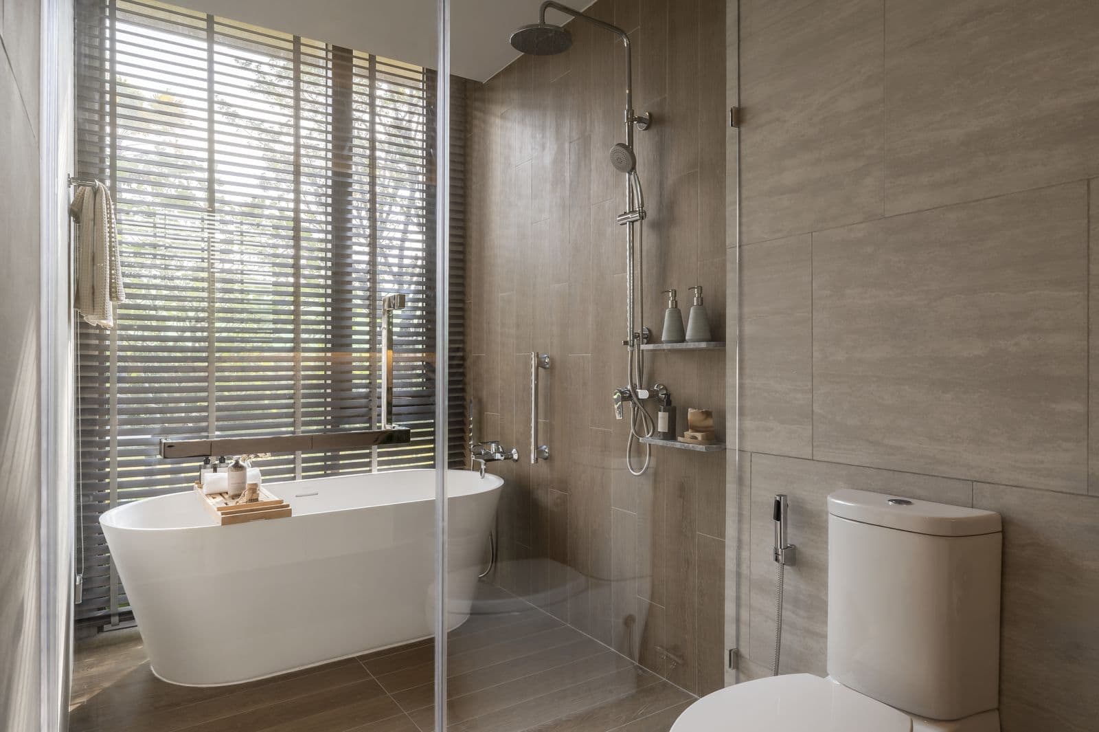 The main bathroom of the finished two-bedroom show unit at PEYLAA Phuket: a freestanding bathtub by a window with blinds, a glass shower and a WC