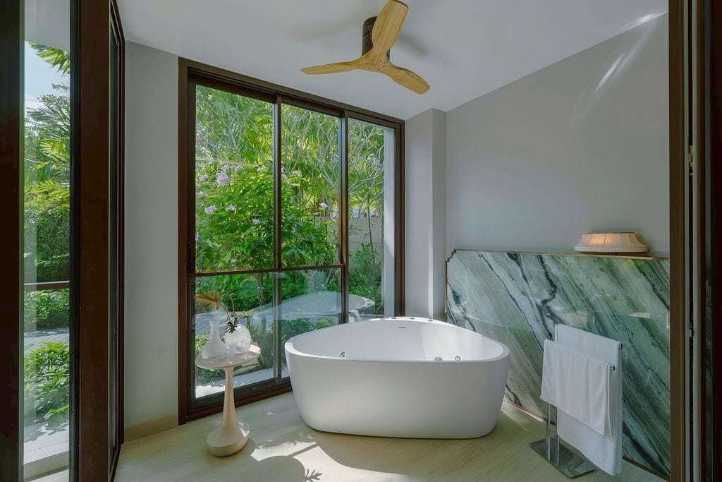 Whirlpool bath on the balcony of the furnished one-bedroom show unit of The Residences at InterContinental Phuket Resort, a green marble wall, a ceiling fan and the tropical garden beyond the glass
