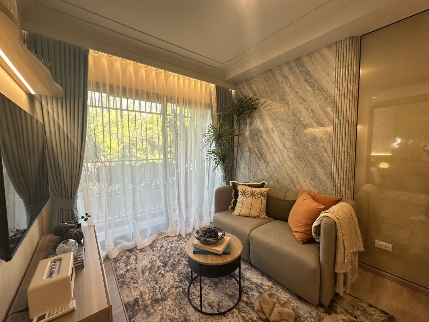 The living room of the furnished one-bedroom show unit at Rhea by Sansiri, Surin, Phuket: a sofa with orange cushions, a round coffee table, a marble-look wall and a window onto trees