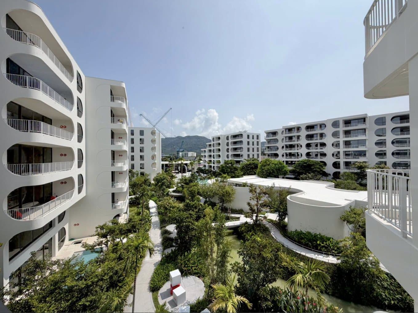 The finished white buildings of The Standard Residences, Phuket Bang Tao around their garden courtyard, with the curved balconies, the pool pavilion, the lagoon and the hills of Phuket behind, photographed at the opening in October 2026