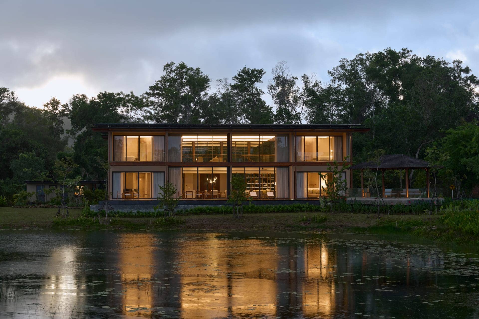 The finished Vana 4 show villa at Tri Vananda, Phuket, at dusk: a two-storey wooden villa lit from inside, reflected in the lake in front of it