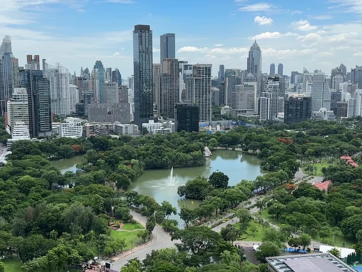 曼谷市中心的 Lumphini Park:樹叢間的湖泊與噴泉,遠處是城市天際線;Embark Estate 拍攝