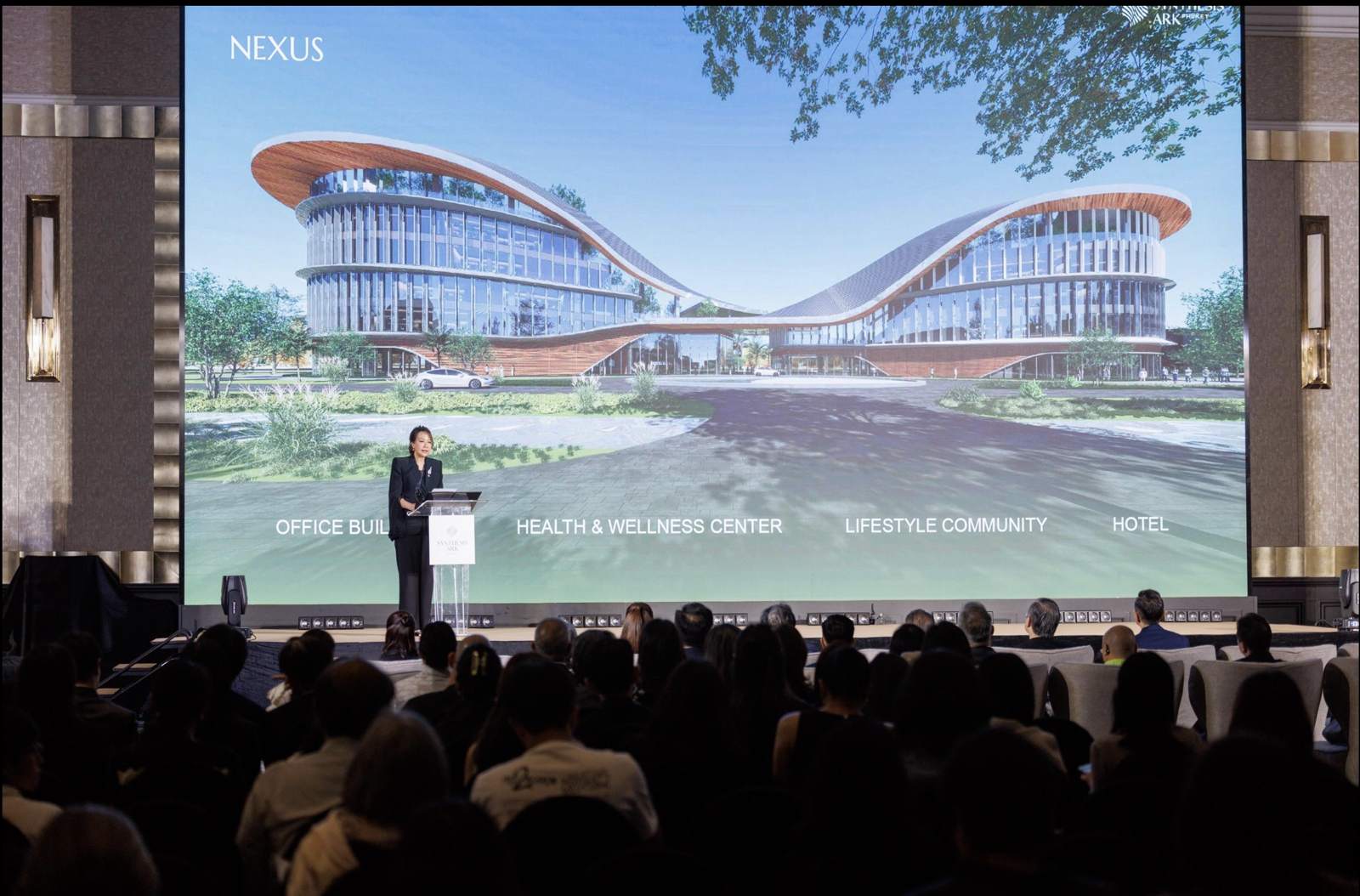Synthesis Ark Phuket 的推介发布会:讲者立于讲台,身后屏幕显示 Nexus 各座建筑,并标示写字楼、健康与养生中心、生活社区与酒店