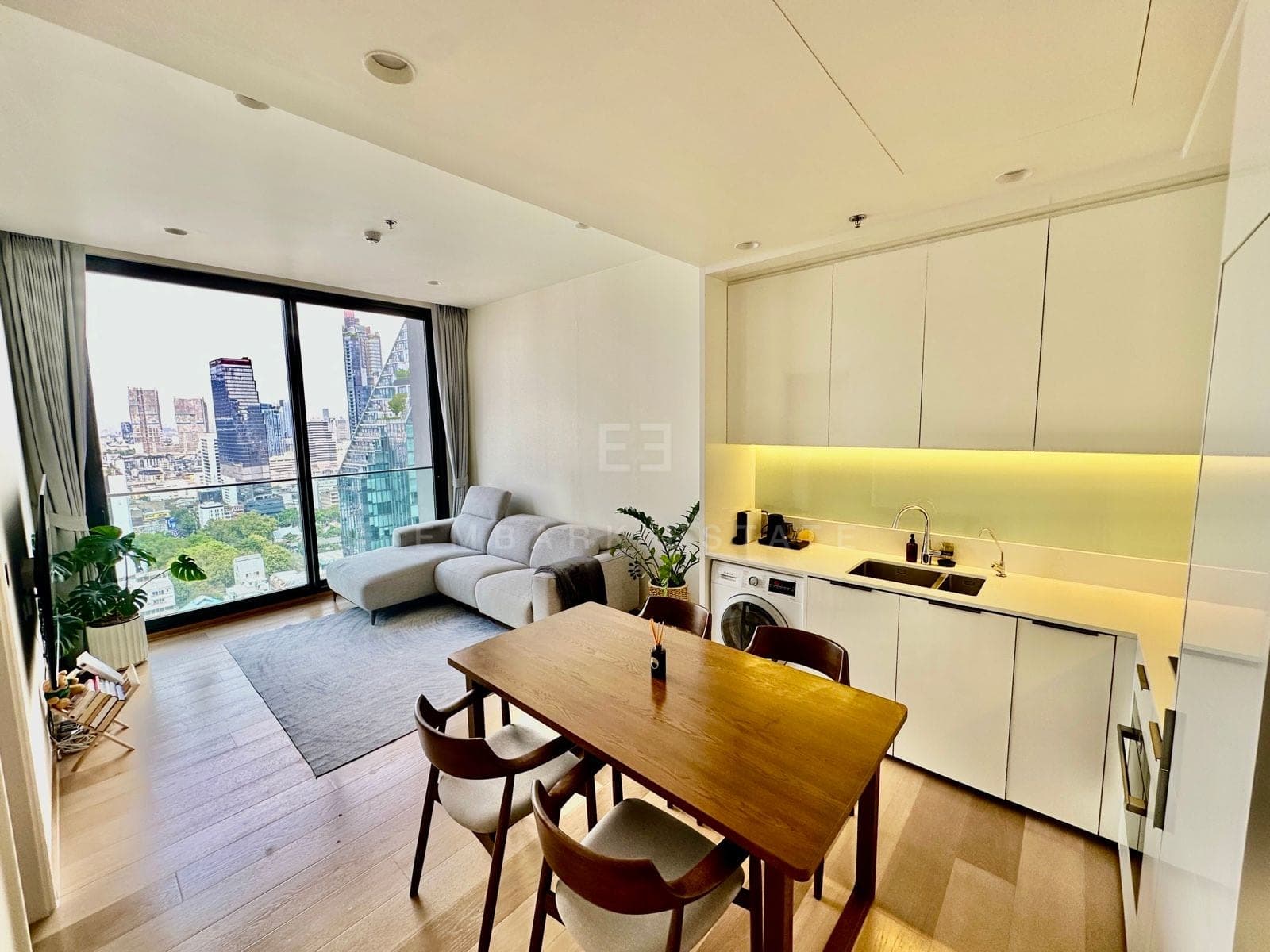 ห้องนั่งเล่นและห้องอาหารแบบเปิดโล่งของยูนิตหนึ่งห้องนอนที่เปิดขายที่ ANIL Sathorn 12 กรุงเทพฯ พร้อมกระจกสูงจรดเพดานมองเห็นเส้นขอบฟ้าเมือง