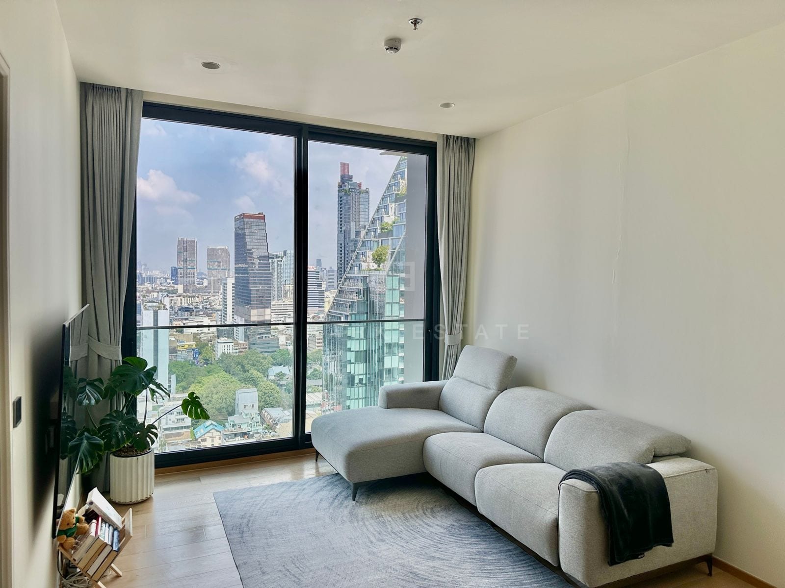 The living room of a 1-bedroom residence at ANIL Sathorn 12, Bangkok, with a grey sofa beside a floor-to-ceiling window framing King Power MahaNakhon and the city skyline