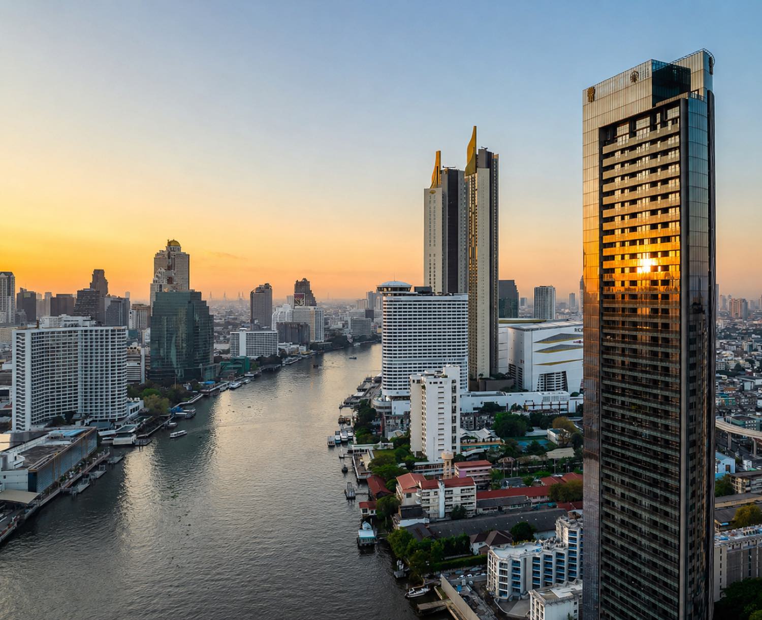 Banyan Tree Residences Riverside Bangkok: the tower on the right lit gold at sunset above the Chao Phraya River, with the Bangkok skyline along the far bank
