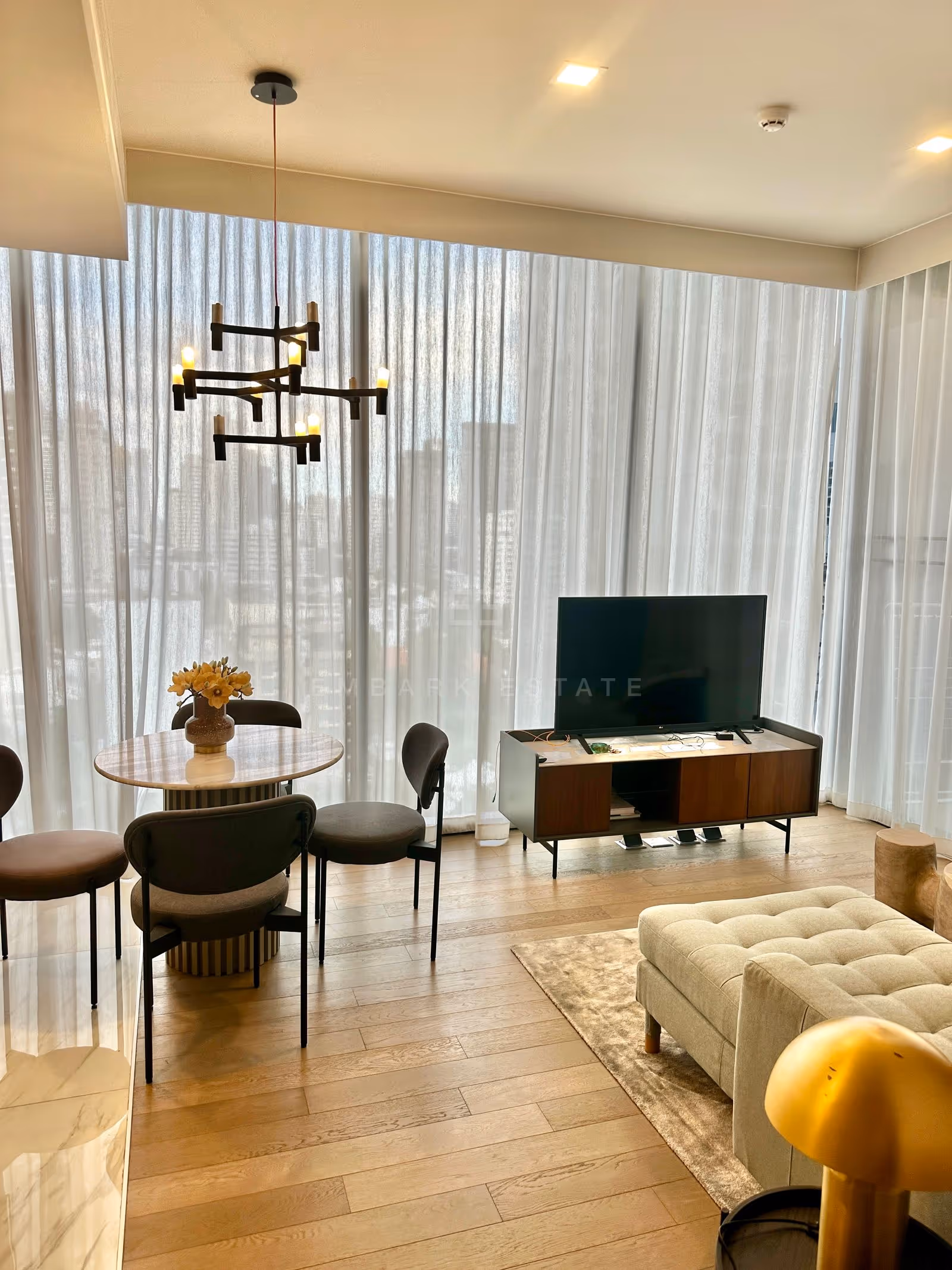 Dining table and television corner of a two-bedroom residence at Celes Asoke, Bangkok, set against full-height sheer-curtained windows with the city skyline beyond.