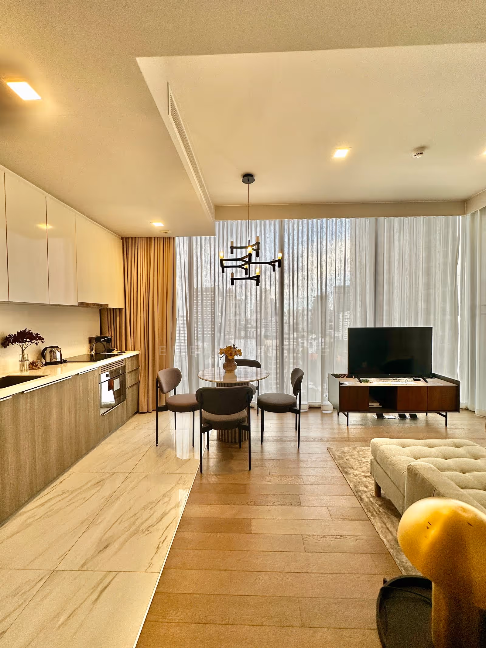 The kitchen run, round dining table and living room of a two-bedroom residence at Celes Asoke, Bangkok, lit by floor-to-ceiling windows along the far wall.