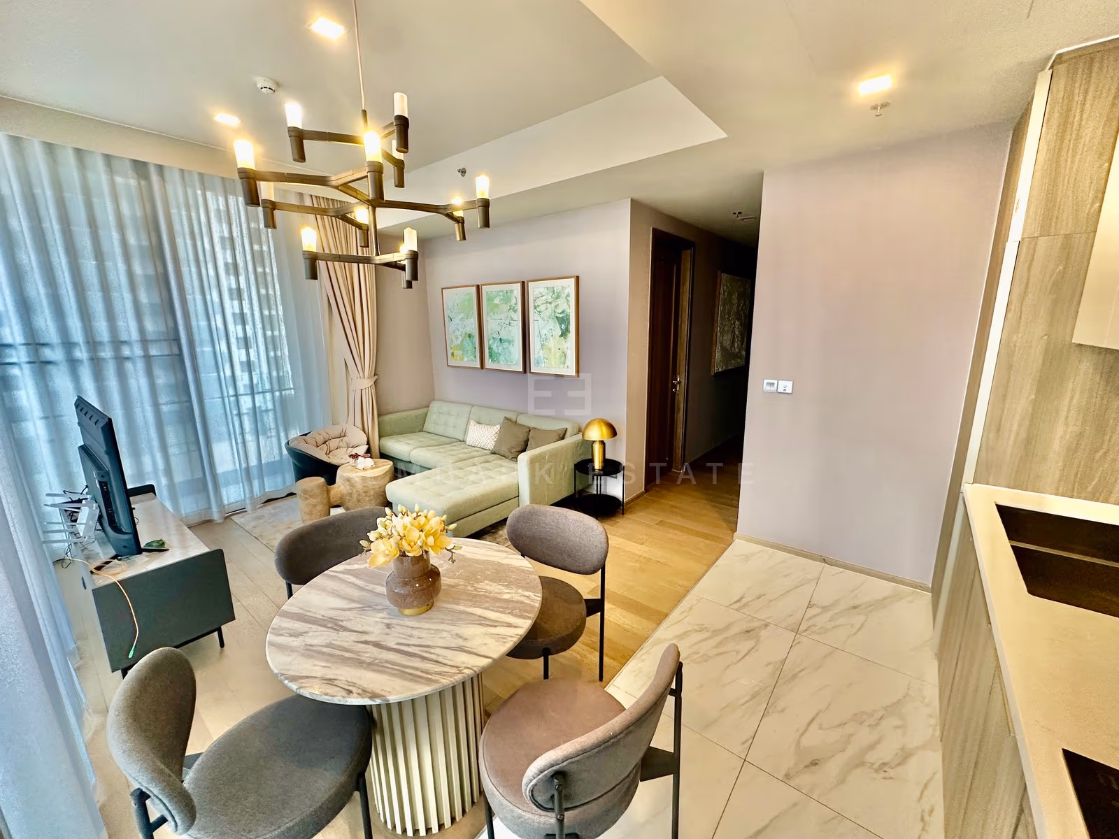Dining area of a two-bedroom residence at Celes Asoke, Bangkok, with a round marble table, four upholstered chairs, a brass chandelier and a pale green sectional sofa beyond.