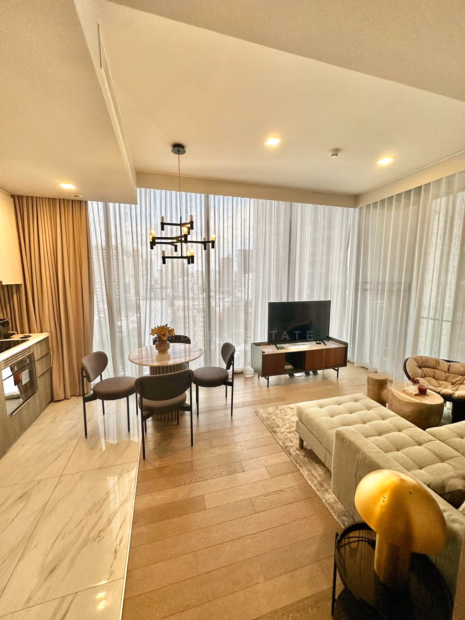 Living room of a two-bedroom residence at Celes Asoke, Bangkok, with floor-to-ceiling windows over the city, a round marble dining table under a brass chandelier and a television console on pale timber floors.