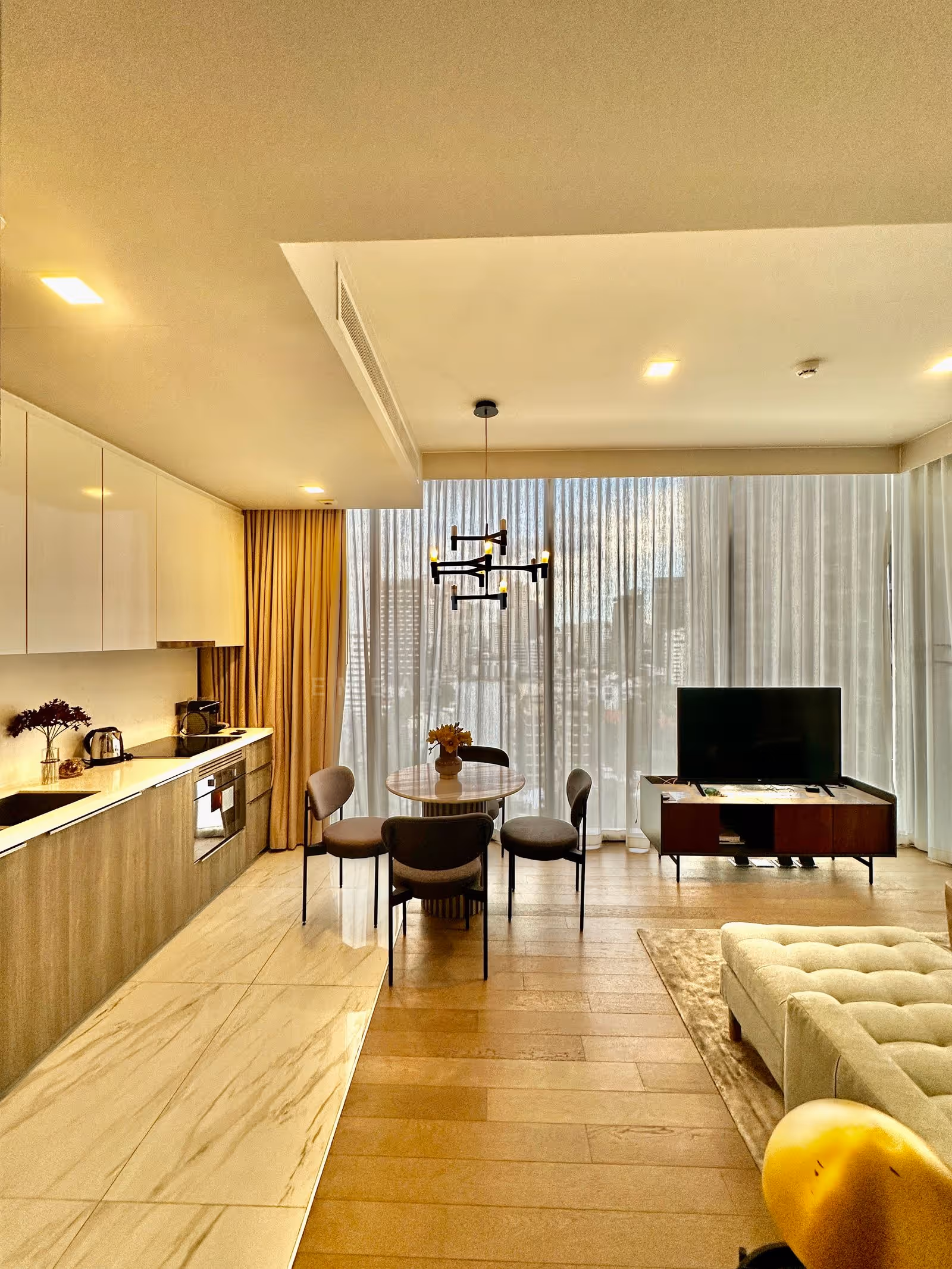 Open-plan kitchen, dining and living room of a two-bedroom residence at Celes Asoke, Bangkok, with white lacquered cabinets, a marble-tiled kitchen floor and full-height windows over the city.