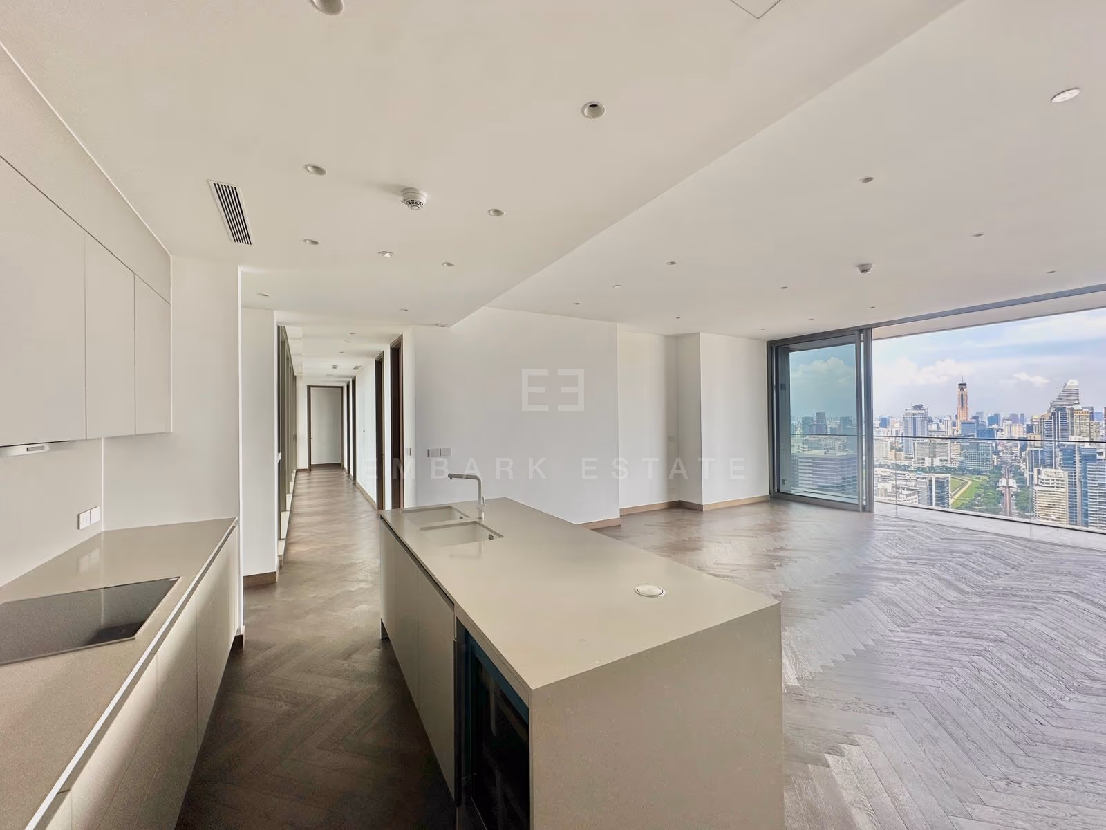 ครัวและห้องนั่งเล่นแบบเปิดของยูนิต 3903 ที่ Dusit Residences, Dusit Central Park กรุงเทพฯ ชั้น 39 เคาน์เตอร์ไอส์แลนด์หินและพื้นไม้โอ๊กลายก้างปลา หน้ากระจกสูงเต็มผนังมองเห็นสวนลุมพินีและเมือง