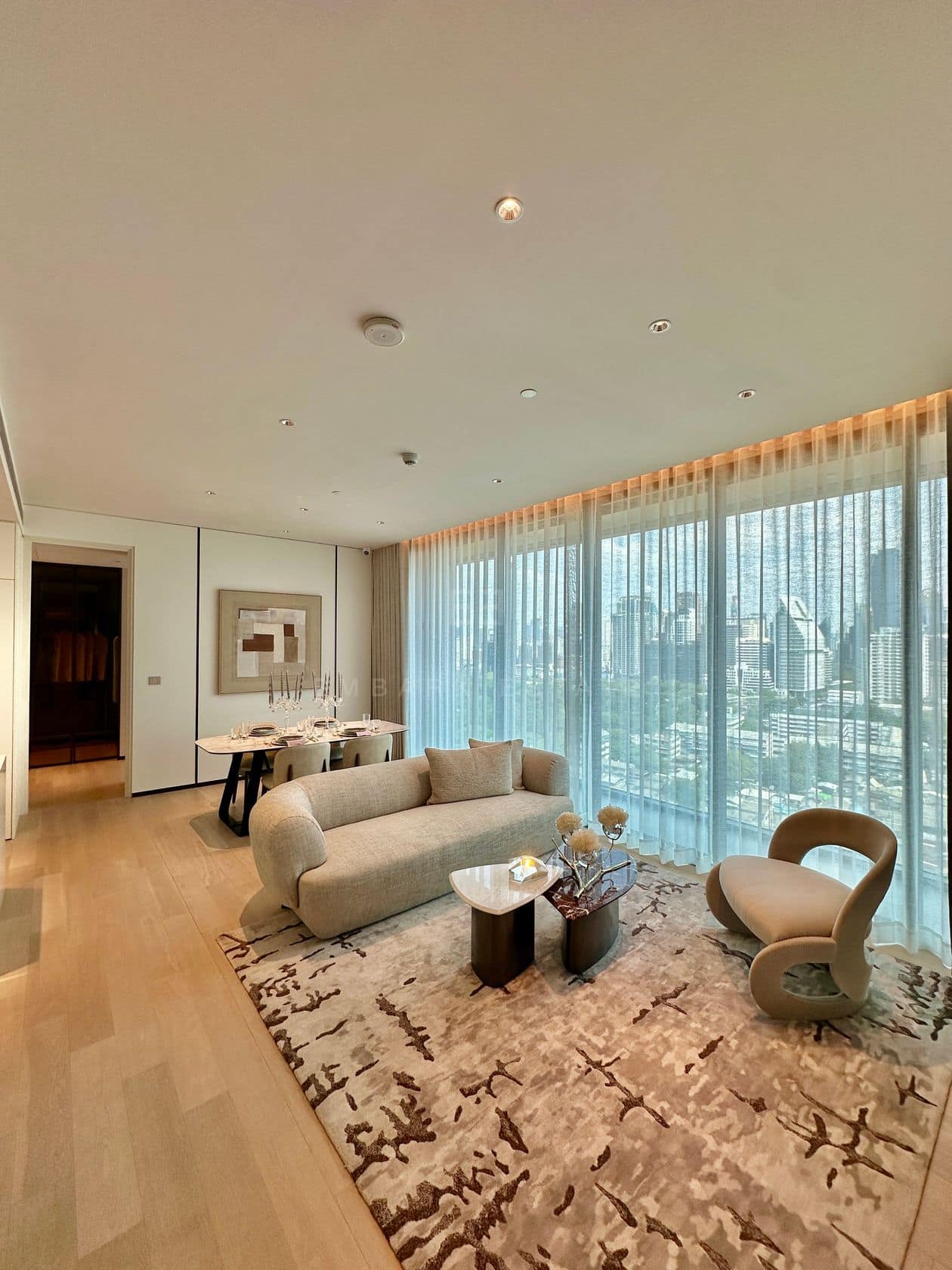 The living room of a two-bedroom residence at EI8HTEEN SEVEN, One Bangkok, with a curved sofa, a lounge chair and floor-to-ceiling views over the central Bangkok skyline