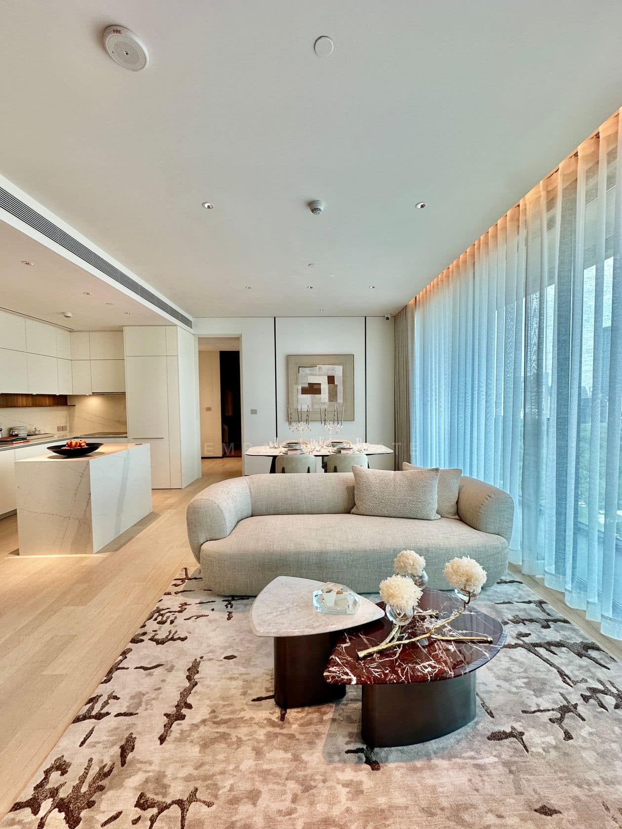 The living room of a two-bedroom residence at EI8HTEEN SEVEN, One Bangkok, with a curved sofa and a marble-topped table, the open kitchen beyond