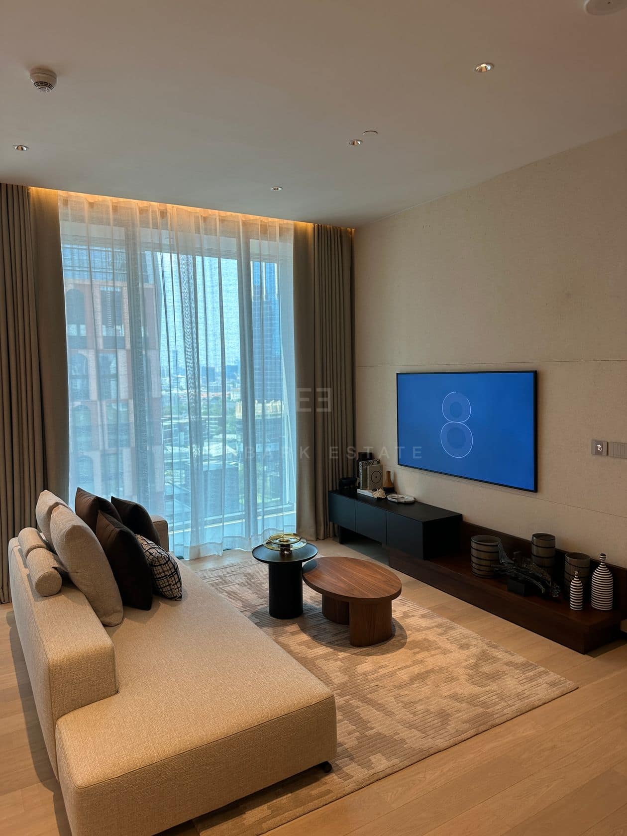 The living room of a one-bedroom residence at EI8HTEEN SEVEN, One Bangkok, with a sofa facing the media wall and floor-to-ceiling city views