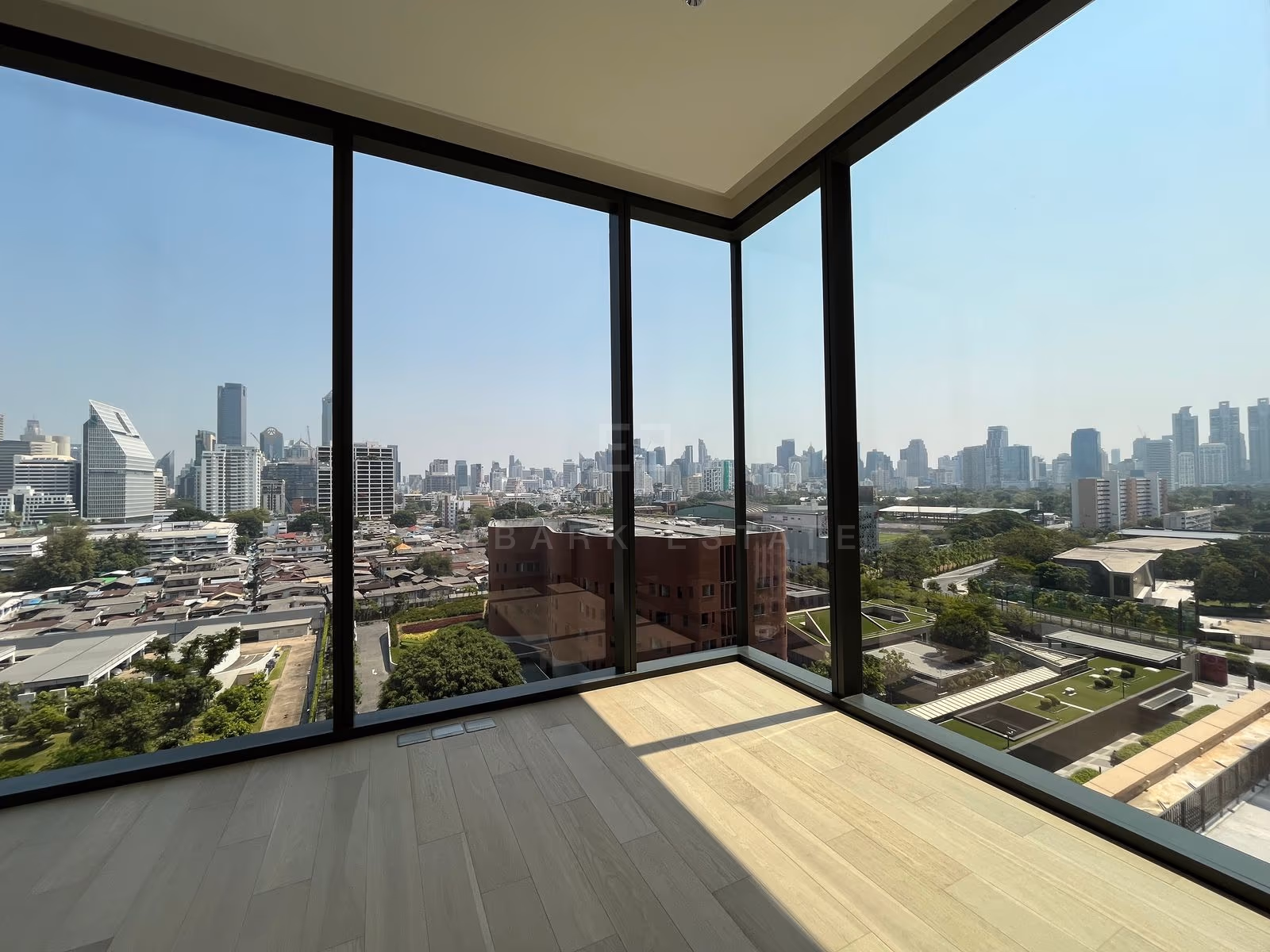 The unfurnished corner bedroom of a one-bedroom residence at EI8HTEEN SEVEN, with floor-to-ceiling glass on two sides overlooking low-rise rooftops, a curved red-brick building and the Bangkok skyline.