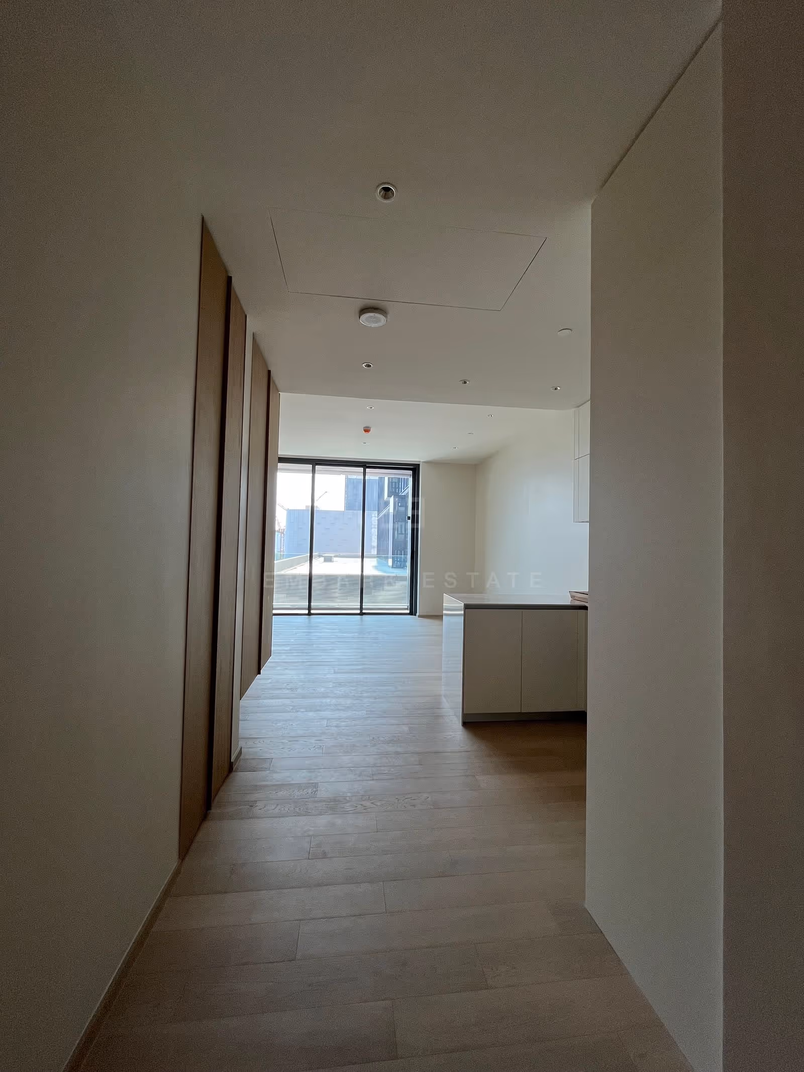 The entrance hall of a one-bedroom residence at EI8HTEEN SEVEN, lined with tall wood-veneer cabinets and leading past the kitchen counter to the living room and balcony door.