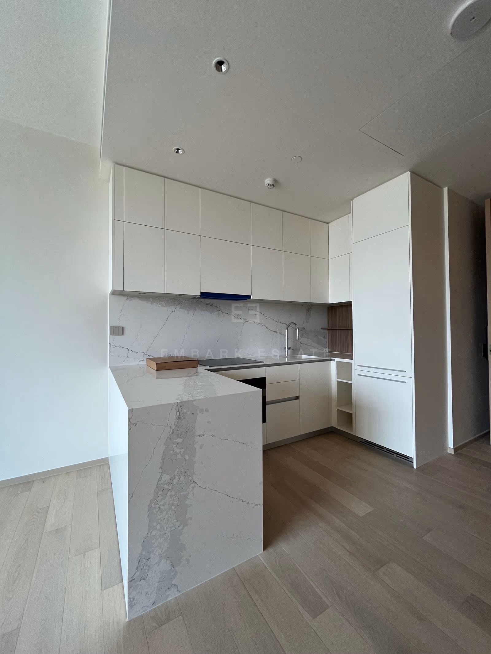 The kitchen of a one-bedroom residence at EI8HTEEN SEVEN, with white handleless cabinets, a marble-look slab backsplash, a waterfall-edge peninsula, a gooseneck tap and a built-in oven.