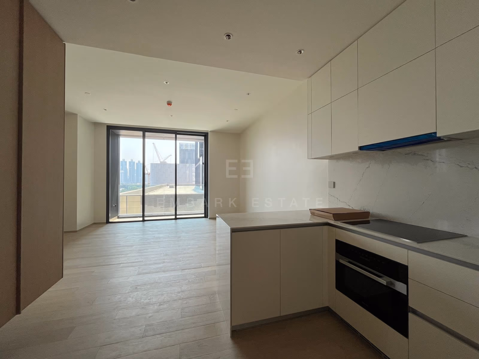 The unfurnished living room of a one-bedroom residence at EI8HTEEN SEVEN, with light oak floors and a sliding glass door to the balcony, beside an open kitchen with a built-in oven and hob.