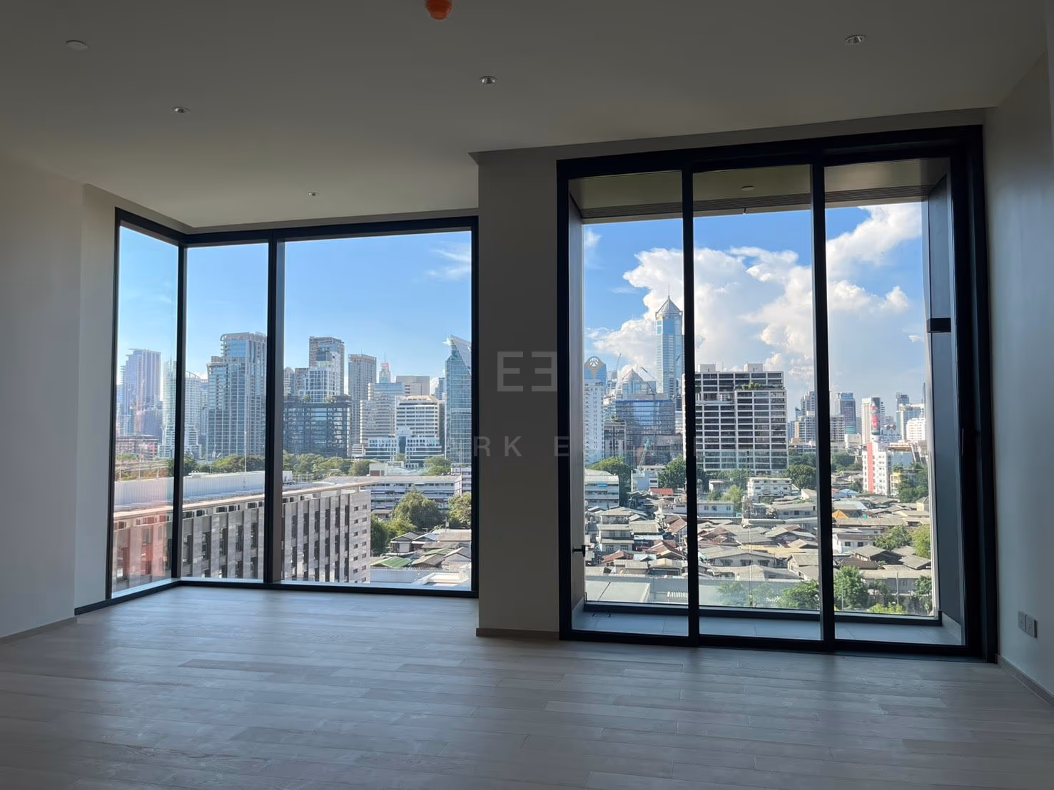 ห้องนั่งเล่นของห้องชุด 1 ห้องนอนที่ EI8HTEEN SEVEN กรุงเทพฯ ในสภาพห้องว่าง ด้านซ้ายเป็นกระจกสูงจรดเพดานแบบเข้ามุม ด้านขวาเป็นประตูกระจกบานเลื่อน เปิดออกสู่วิวหลังคาบ้านเรือนและตึกสูงไกลออกไป