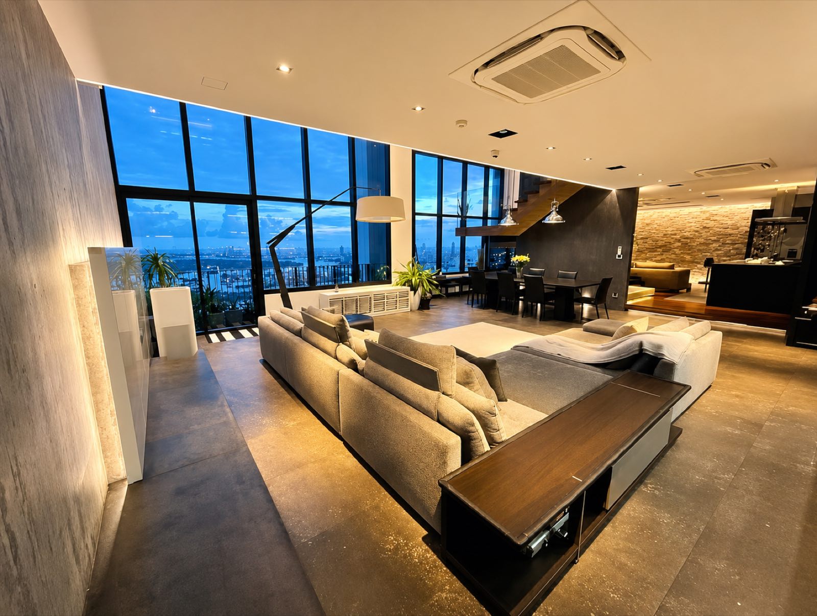 The double-height living and dining room of a duplex penthouse at The Emporio Place, Sukhumvit 24, with floor-to-ceiling glazing and the Bangkok skyline at dusk