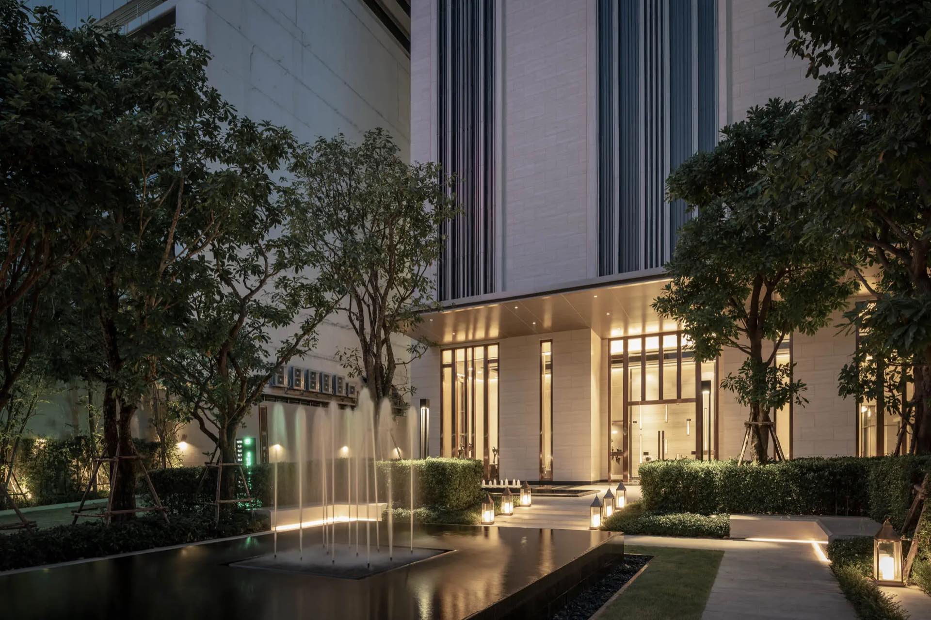 The ESSE Asoke: Illuminated tower-base entrance with a black reflecting pool and water jets in the evening garden