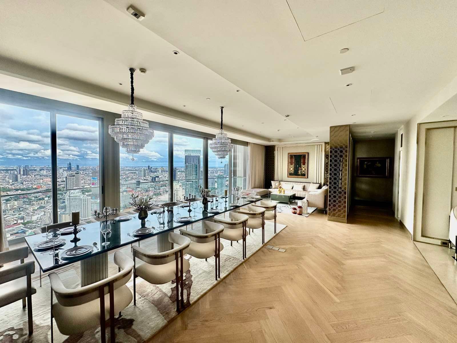 Open-plan dining and living space with a long glass dining table set beneath two crystal chandeliers, a sofa seating area beyond, herringbone wood floor, and floor-to-ceiling windows looking over the city, Mandarin Oriental Residences Bangkok.
