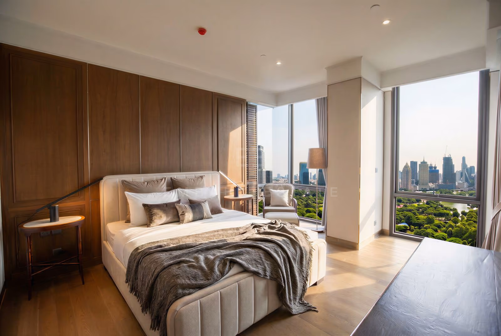 Bedroom of the 25th-floor penthouse at MUNIQ Langsuan, Bangkok, with a cream upholstered bed, a timber-panelled wall and corner floor-to-ceiling windows over the treetops of Lumphini Park and the city skyline.