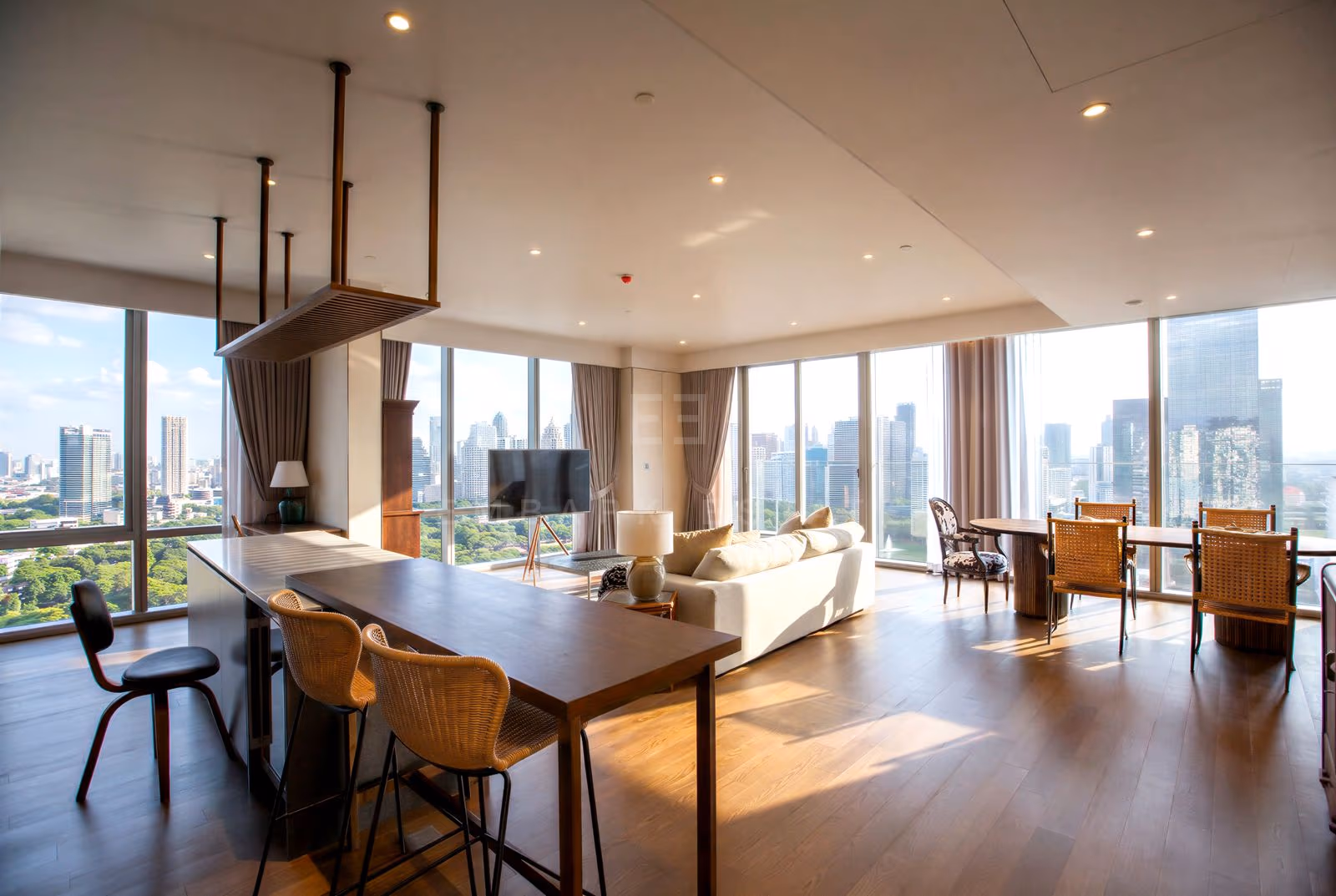 Open-plan living and dining area of the penthouse at MUNIQ Langsuan, Bangkok, with a cream sofa, a long timber bar table with rattan stools, a dining table by the window and floor-to-ceiling glass over treetops and towers.