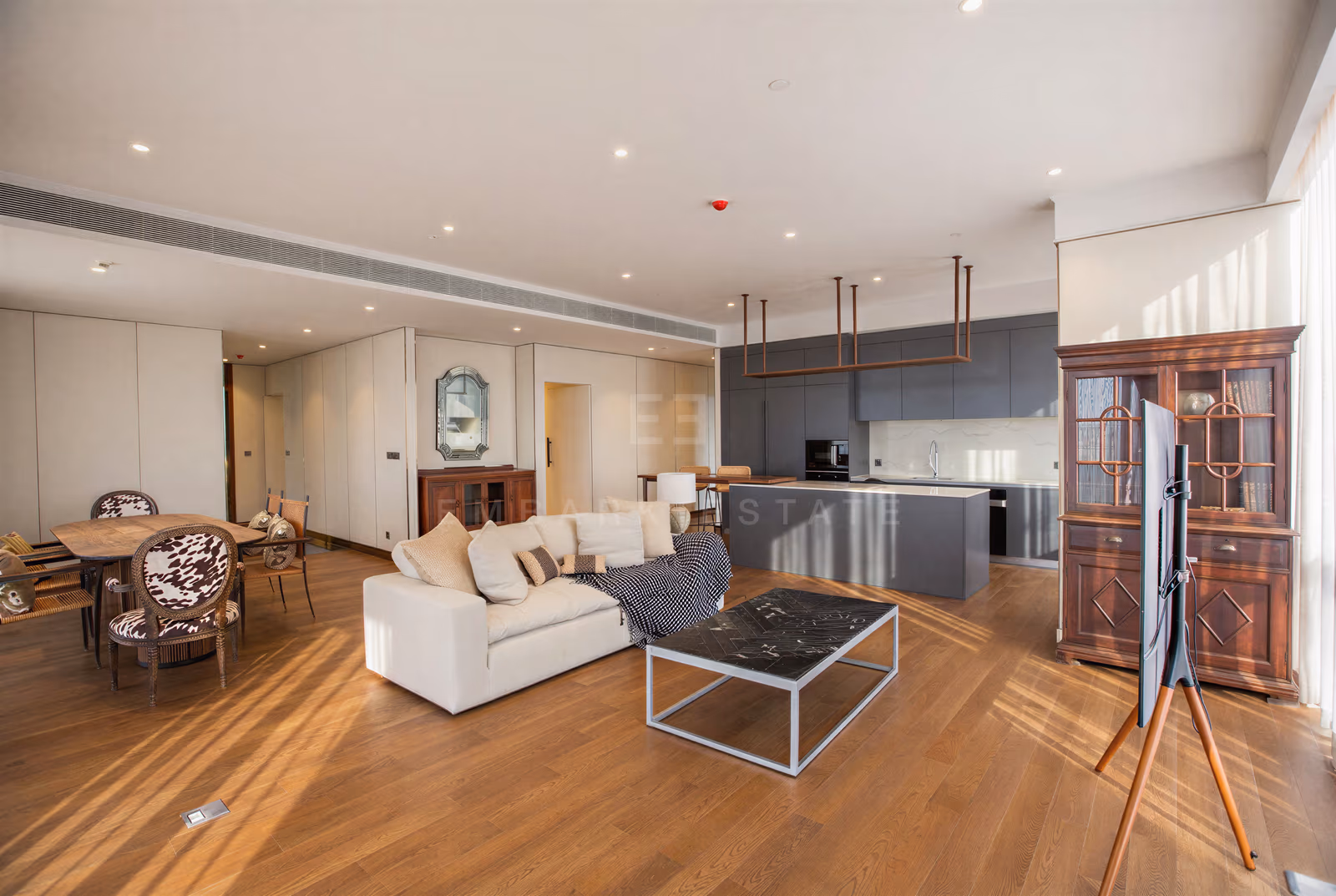 Living area and kitchen of the penthouse at MUNIQ Langsuan, Bangkok, with a cream sofa facing a black marble coffee table, a grey island kitchen with a built-in oven behind it and timber flooring.