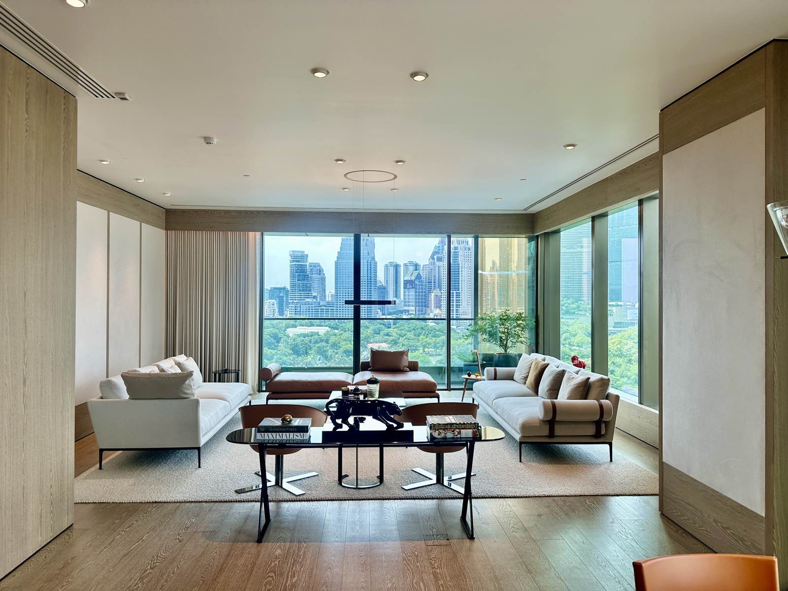 Living room of a residence at NIMIT Lumphini Park North in Bangkok, pale oak panelling and floor-to-ceiling glass on two sides, with Lumphini Park and the Sathorn skyline beyond, photographed October 2026