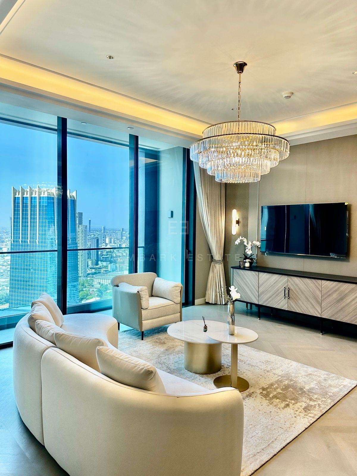 The living room of a two-bedroom residence at ONE89 Wireless, One Bangkok, with a curved sofa, a crystal chandelier and floor-to-ceiling views over the central Bangkok skyline