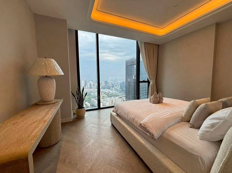 Principal bedroom of a two-bedroom residence at ONE89 Wireless, with a cream upholstered bed, an oak console with a table lamp and floor-to-ceiling windows looking over the city.