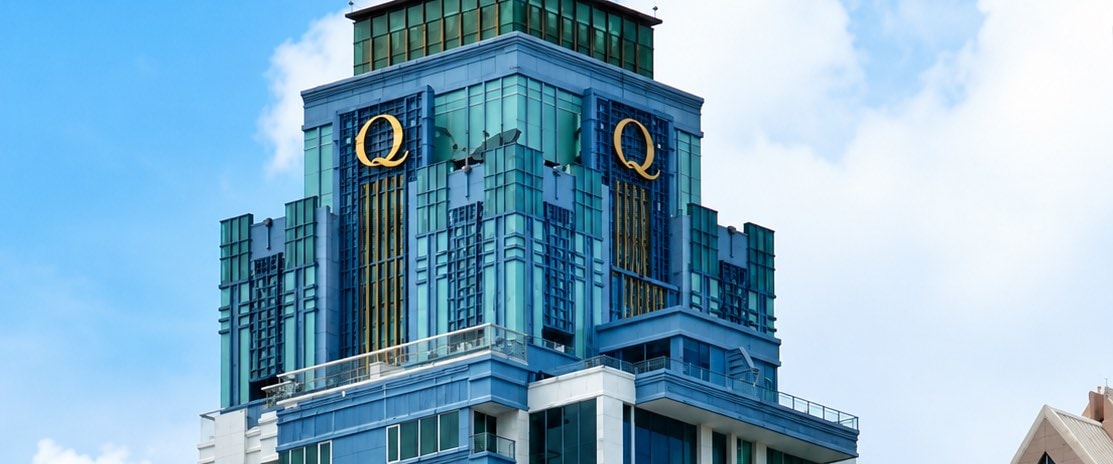 The blue-glass crown of the Q Langsuan tower with its two gold Q emblems, above the Langsuan neighbourhood in central Bangkok
