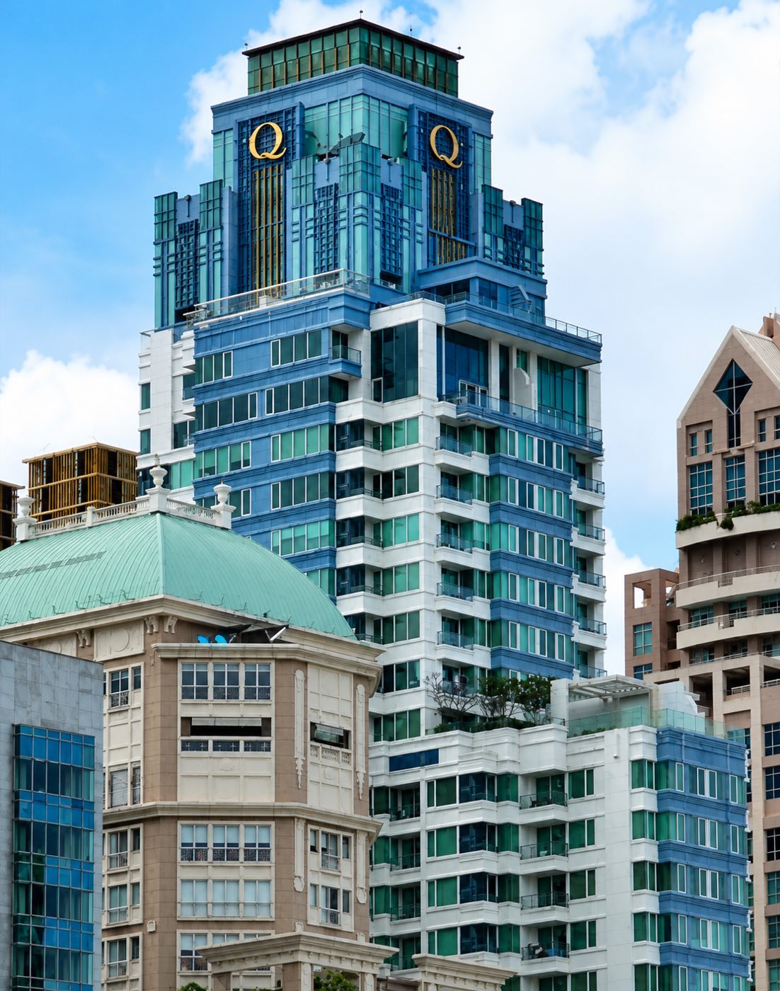 The Q Langsuan tower with its gold Q emblems on the blue-glass crown, above the Langsuan neighbourhood in central Bangkok