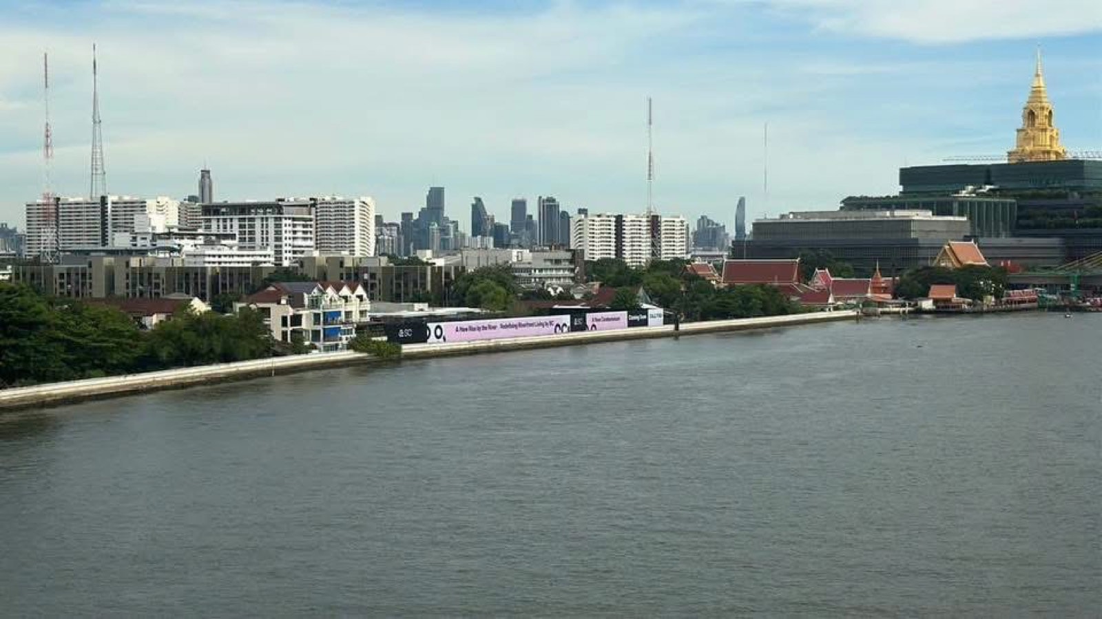 ที่ตั้งโครงการ Reference BAIE River Bangpho ถ่ายจากแม่น้ำเจ้าพระยา กำแพงริมน้ำสีขาวพร้อมป้ายโครงการของ SC Asset ย่านบางโพอาคารเตี้ยด้านหลัง และอาคารรัฐสภายอดทองอยู่ฝั่งตรงข้ามทางขวา