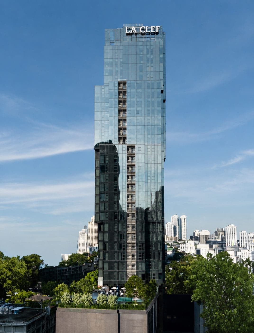 The Residences 38: the faceted glass tower topped by the LA CLEF sign, above the trees and the Thonglor skyline in central Bangkok