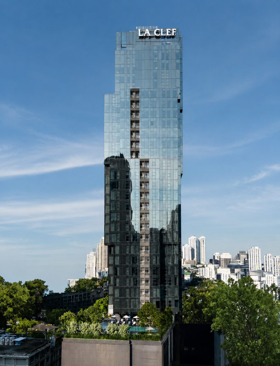 The Residences 38: the faceted glass tower topped by the LA CLEF sign, above the trees and the Thonglor skyline in central Bangkok